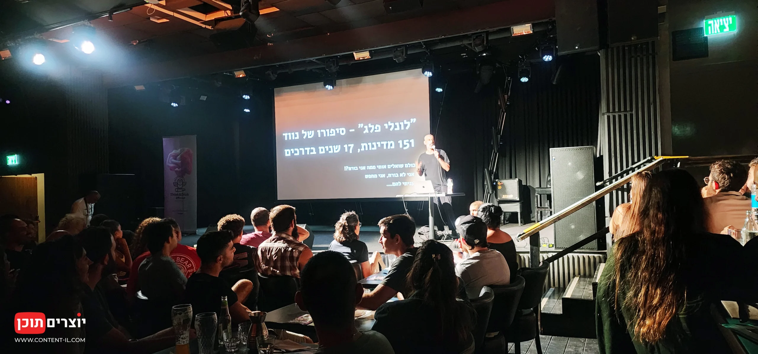 לונלי פלג עומד על הבמה באולם גריי בתל אביב בפתיחת ההרצאה, מול קהל מלא